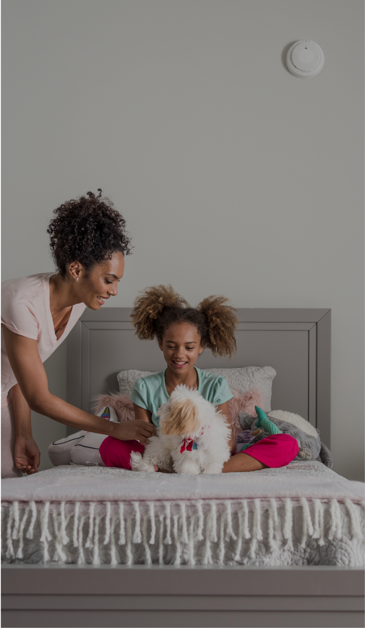 Mom tucking her child into bed with an ADT smoke detector mounted on a wall alongside a door/window sensor on a window